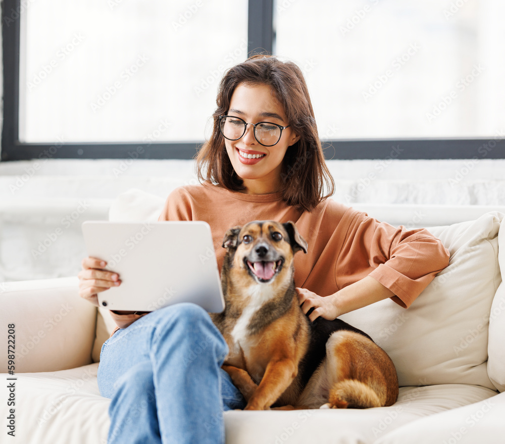 Happy young woman spending time with her pet dog at home sitting on couch and browsing in tablet. Happy young woman spending time with her pet dog at home sitting on couch and browsing in tablet.
