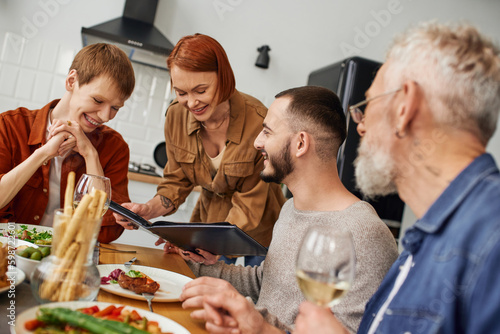 smiling mother showing photo album to son next to his gay boyfriend during family supper at home. 