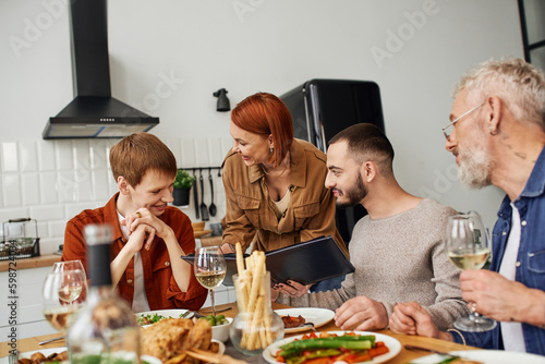 cheerful redhead woman showing family photo album to smiling son with gay boyfriend near delicious supper served in kitchen. 