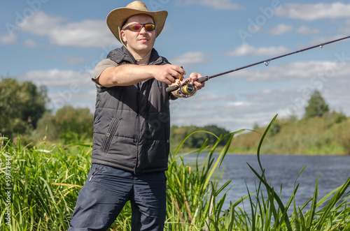 Fisherman in a hat and glasses on the river bank with a spinning rod in his hands.