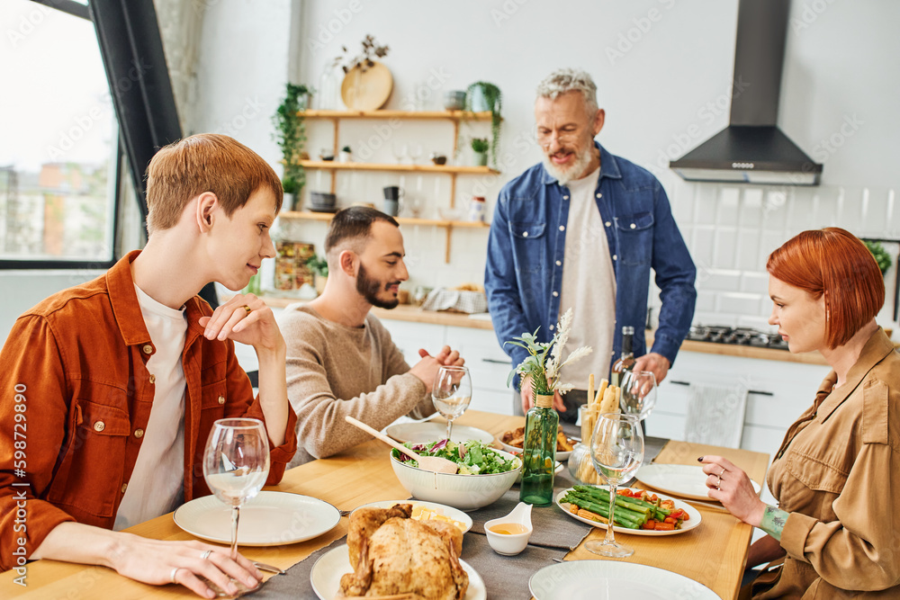 bearded man standing with bottle of wine near son with gay partner during family dinner in kitchen. bearded man standing with bottle of wine near son with gay partner during family dinner in kitchen.