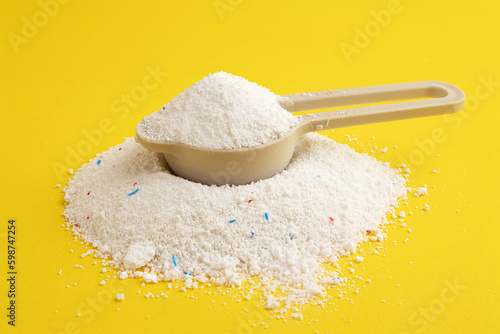 A pile of washing powder and a measuring spoon on a yellow background. Powder with blue and red granules.