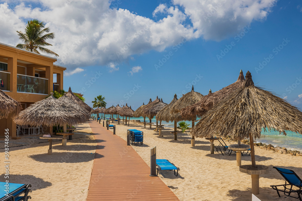 Beautiful view of wooden path between hotel buildings and sun umbrellas on beach along coastline. Aruba. 