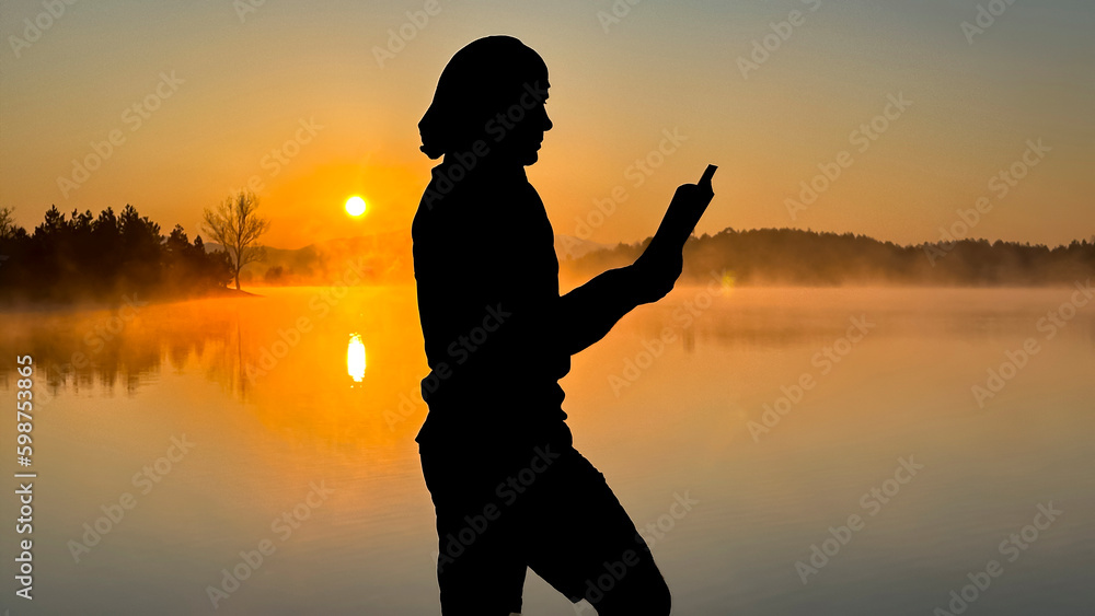 Fototapeta premium silhouette of person reading a book by the lake at sunrise