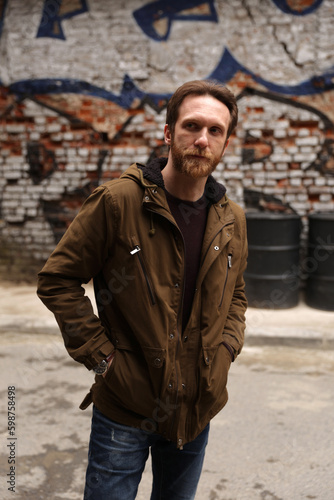 Bearded caucasian man with brown jacket posing in front of a graffiti wall