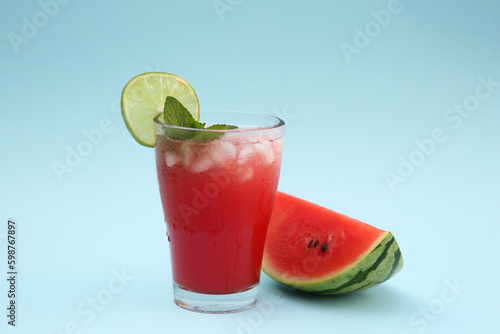 Glass of delicious drink with lime, ice cubes and cut fresh watermelon on light blue background