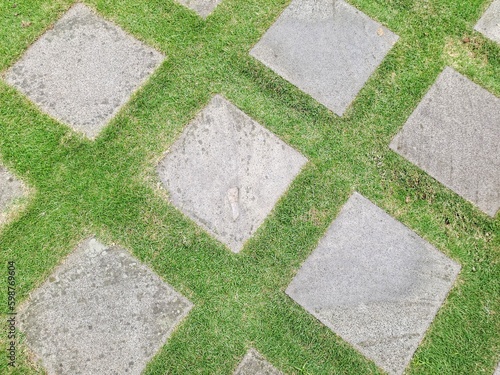Pattern of square cement floor tiles with green . walkway with green grass in the park. Stone pathway and green grass at park. Garden Stepping Stone Path through Green Grass Law.