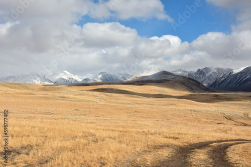 Field roads cross high hills in the dry autumn steppe at the foot of high snow-capped mountains.