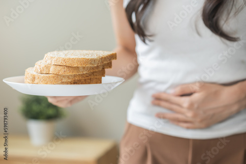Gluten allergy, asian young woman hand holding, refusing to eat white bread slice on plate in breakfast food meal at home, girl having a stomach ache. Gluten intolerant and Gluten free diet concept. photo