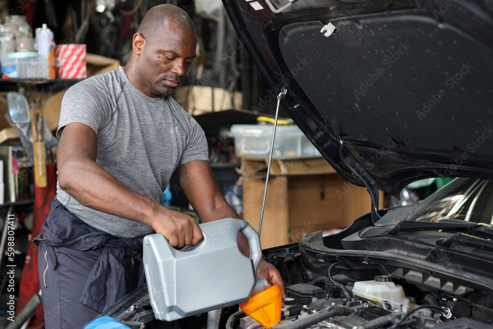 Man car mechanic or worker in half uniform pouring oil into car and fixing breakdown vehicle at garage station. Concept of car center repair service and energy fuel concept. Man car mechanic or worker in half uniform pouring oil into car and fixing breakdown vehicle at garage station. Concept of car center repair service and energy fuel concept.