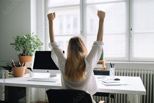 Rear view of a female executive in her home office celebrating success with her arms raised.