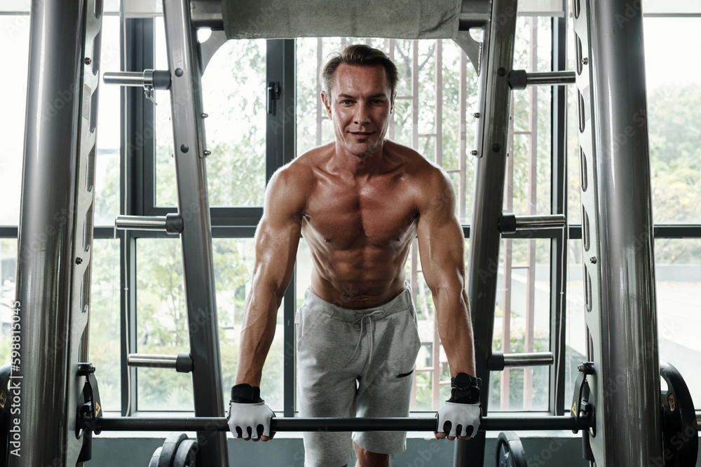 Naklejka premium Portrait of sweaty positive sportsman leaning on barbell and smiling at camera