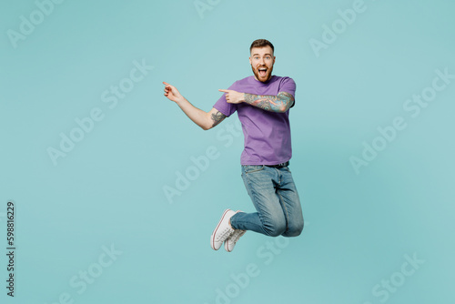 Full body excited young man he wear purple t-shirt jump high point index finger aside indicate on workspace area isolated on plain pastel light blue cyan background studio portrait. Lifestyle concept.