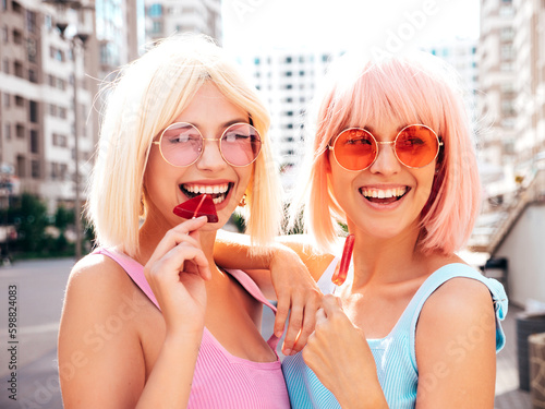 Two young beautiful smiling hipster female in trendy summer clothes. Sexy carefree women posing in the street in wigs. Positive funny models having fun at sunset. In sunglasses. Eating candy lollipop