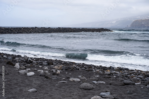 Black sand beach with waves. Breakwater on one side. Cliffs with houses in the background. Stones in the sand. Cloudy day. Puerto de la Cruz, Tenerife, Canary Islands, Spain.