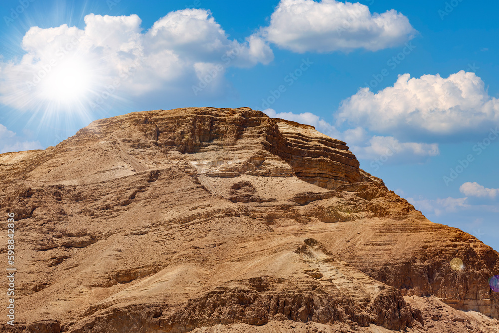 Background of mountains and sun in the blue sky in Dead Sea, Israel. Beautiful desert landscape among the sands and stones. Large stone ridge against the background of a blue sky with white clouds