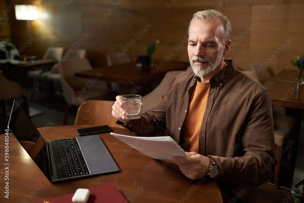 Mature man drinking water and reading contract while working on laptop at table in cafe