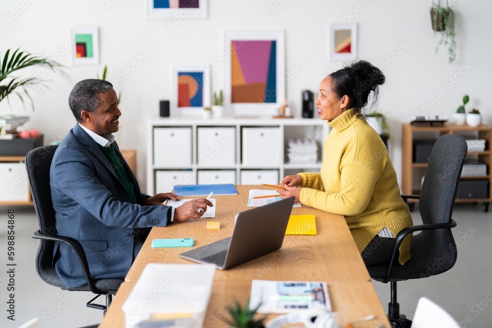 Side view of smiling Ethiopian woman and man in smart casual clothes talking while looking at each other in modern office Side view of smiling Ethiopian woman and man in smart casual clothes talking while looking at each other in modern office