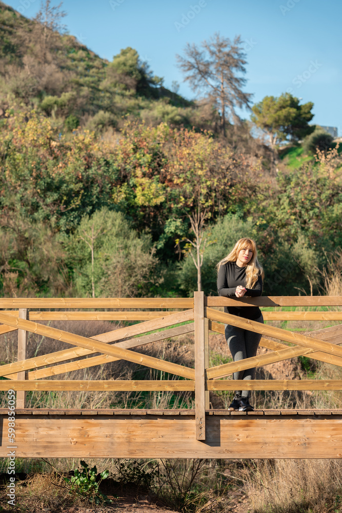 beautiful middle-aged blonde woman in sportswear leaning on the fence of a wooden bridge with green trees in the background