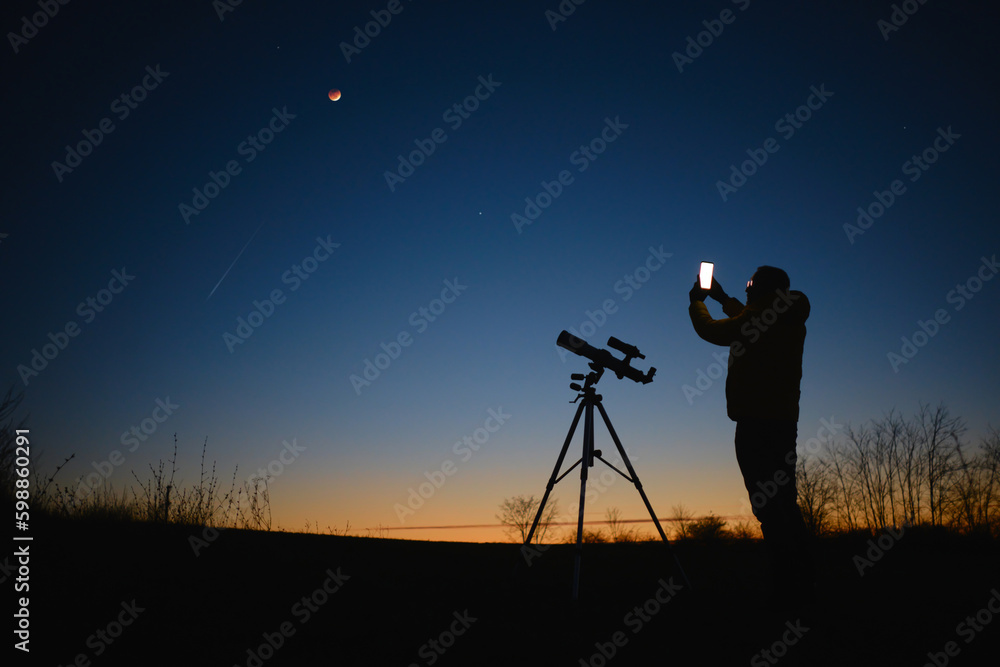 Astronomer looking at the starry skies with planets, falling stars and Moon eclipse with a telescope while using modern smartphone.