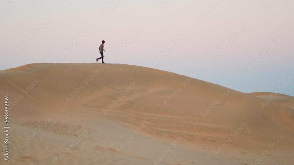 Indian man running down on the sand dunes of the Thar desert at Khuri Sand dunes near Jaisalmer in Rajasthan, India. Tourist running down from top of the sand dune during the early morning. Indian man running down on the sand dunes of the Thar desert at Khuri Sand dunes near Jaisalmer in Rajasthan, India. Tourist running down from top of the sand dune during the early morning.