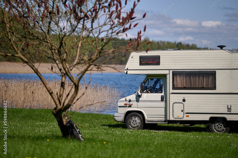 old school white camper parked on the grass by the lake. A vintage house on wheels in a sunny aura.