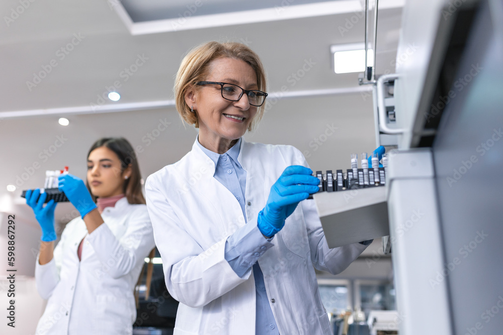 In Laboratory Female Scientist Inspects Medical Equipment Analyzing Test Tube with Blood Samples. Team of Researcher Work in Pharmaceutical Laboratory. In Laboratory Female Scientist Inspects Medical Equipment Analyzing Test Tube with Blood Samples. Team of Researcher Work in Pharmaceutical Laboratory.
