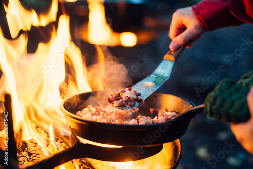 Person frying bacon in pan over campfire