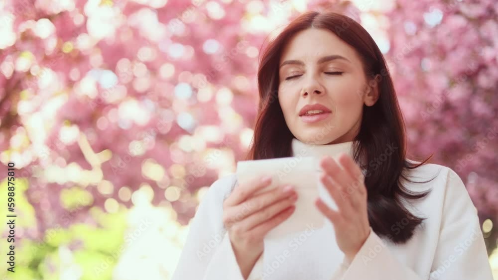 Portrait of allergic unhealthy sick young dark haired woman sneezing in tissue blowing running nose with seasonal allergy symptoms on pink flowers blossom trees background Portrait of allergic unhealthy sick young dark haired woman sneezing in tissue blowing running nose with seasonal allergy symptoms on pink flowers blossom trees background