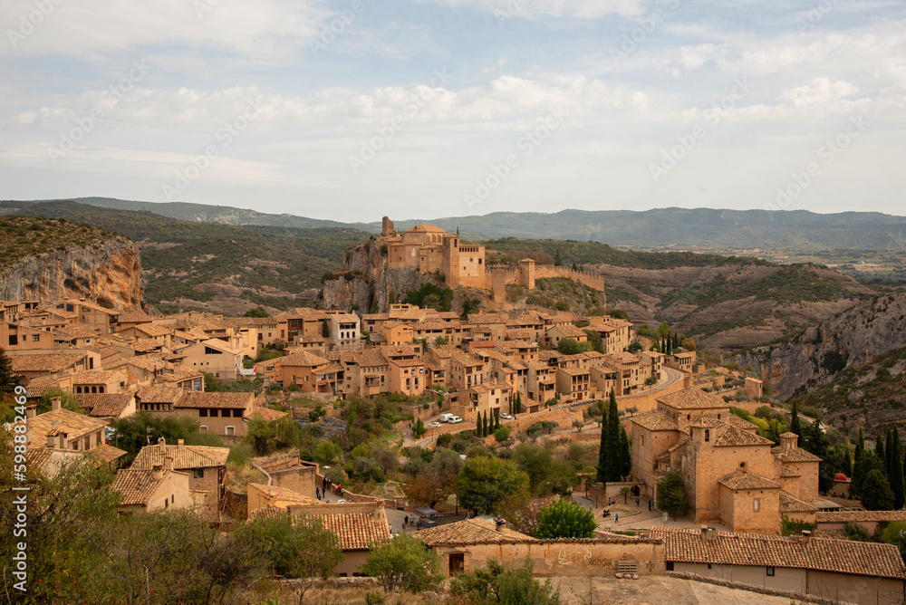 Vista panorámica del turístico pueblo medieval de Alquezar lleno de pequeñas casas de ladrillo y una gran iglesia en la parte superior rodeada por un paisaje montañoso en un día claro.