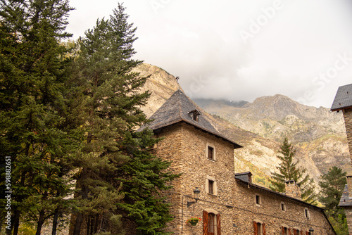 Stone house with slate roofs next to large pine trees and an impressive rocky mountain in the background, mountain scenery in the province of Huesca Aragon on a cloudy day. Benasque.