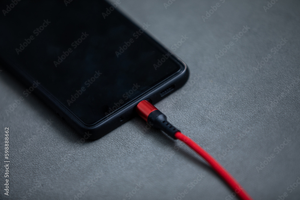 A black smartphone is lying and is connected to the power supply with a red wire. A black smartphone is lying and is connected to the power supply with a red wire.