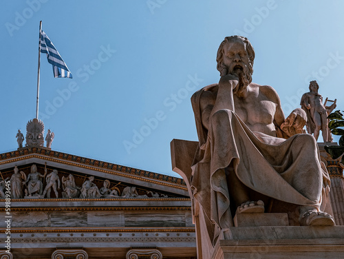 A Greek flag on the academy of Athens building and statues of Socrates, the ancient philosopher and Apollo god. Cultural trips to Athens, Greece.