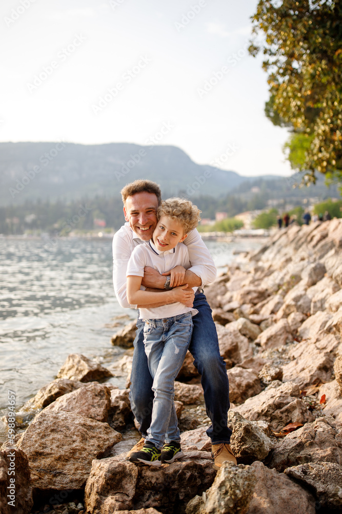 Brunette father hugging and smiling with his blondie curly hair son at the stones of Italian Garda lake 