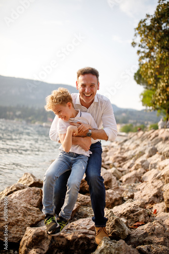 Brunette father and his blondie curly hair son having fun and tickle at the stones of Italian Garda lake 