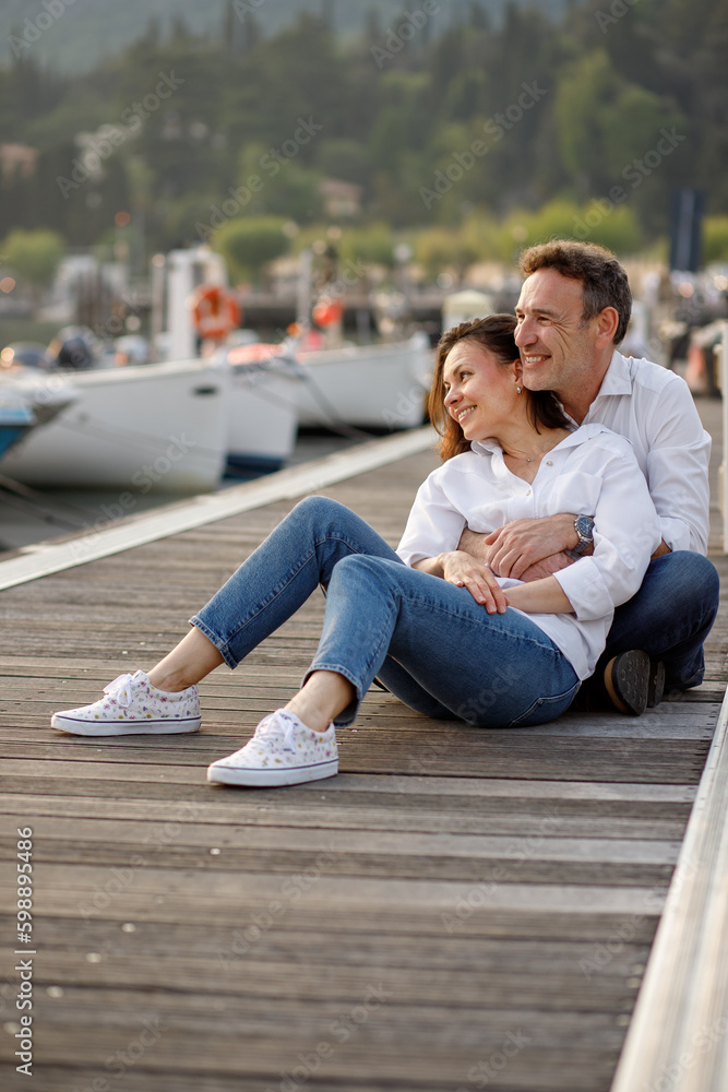 Stylish couple in love in white shirts hugging each other smiling at the wooden pier at Italian Garda lake Stylish couple in love in white shirts hugging each other smiling at the wooden pier at Italian Garda lake