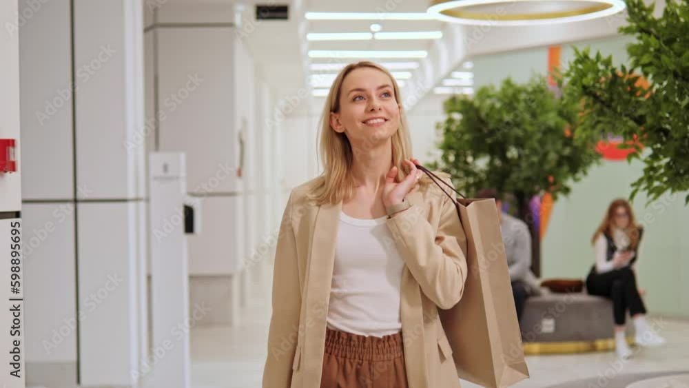 Gorgeous, happy, beautiful, joyful blonde girl holds shopping bags in hands and looking around a shopping center. Woman smiles, enjoys her purchases while people are in the background using gadget. Gorgeous, happy, beautiful, joyful blonde girl holds shopping bags in hands and looking around a shopping center. Woman smiles, enjoys her purchases while people are in the background using gadget.