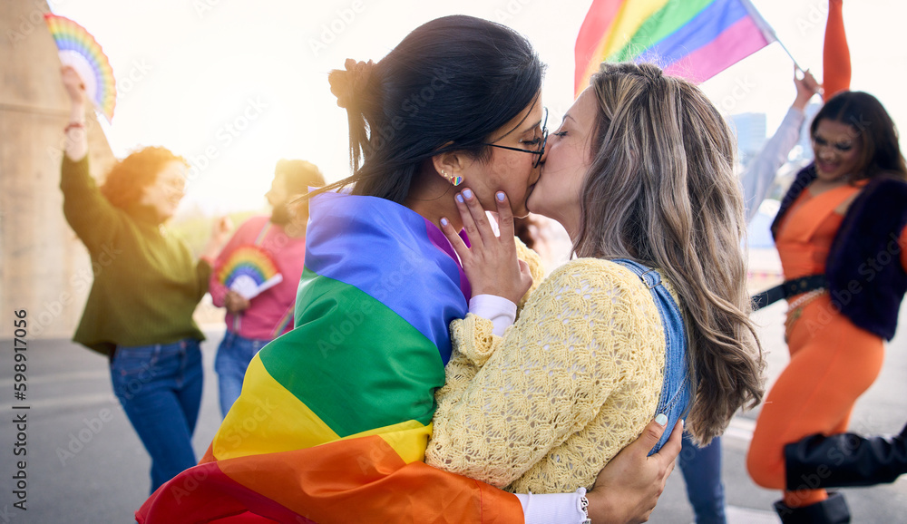 Two young loving girls kissing on gay pride festival day outdoors. Group of friends celebrating lgbt party on background with rainbow flag and fans. Generation z and types of sexuality. - obrazy, fototapety, plakaty 