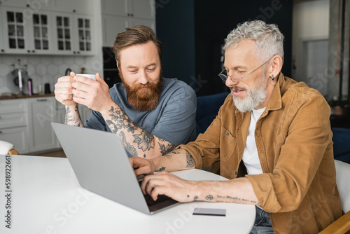 Bild auf Leinwand Tattooed gay couple with coffee using laptop near credit card at home