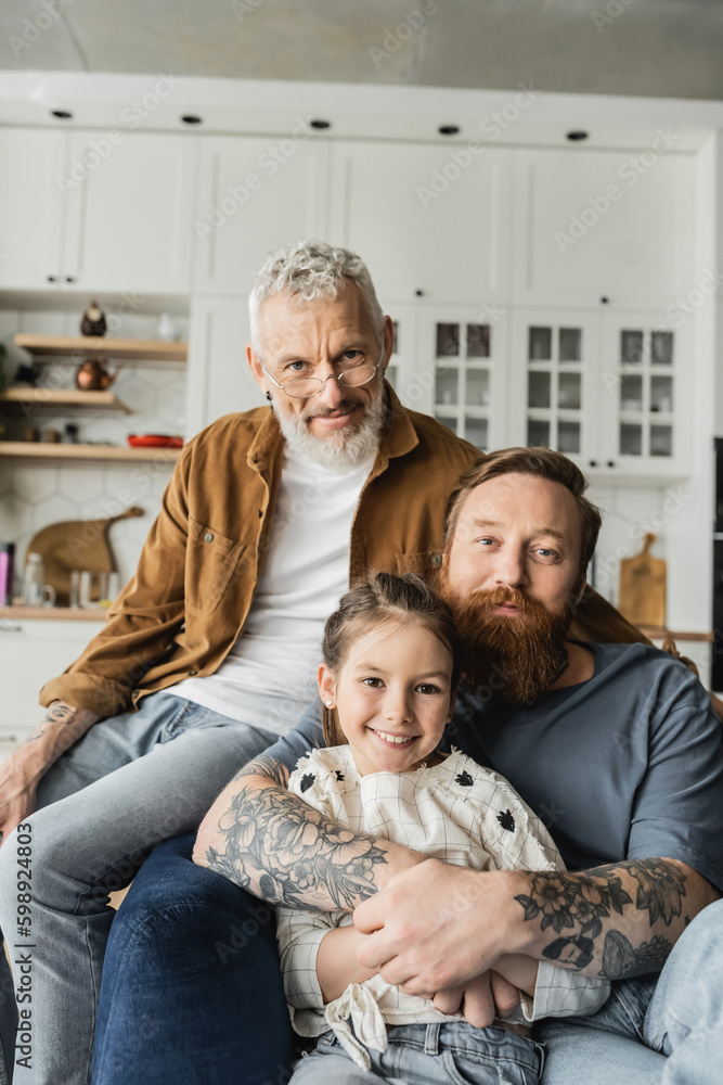 Homosexual man hugging smiling daughter near partner and looking at camera at home. 