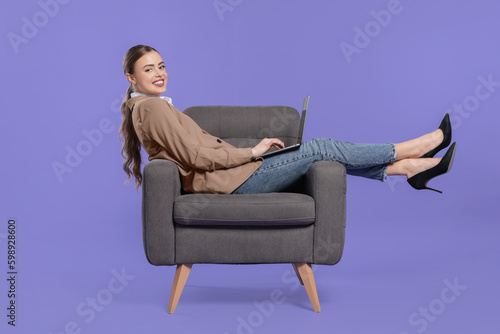 Happy woman with laptop sitting in armchair on violet background © New Africa