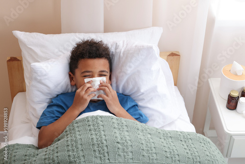 African-American boy with scarf and tissue blowing nose in bed indoors, above view. Cold symptoms