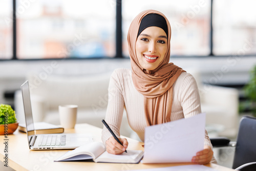 beautiful smiling muslim woman in traditional religious hijab works remotely on laptop from home