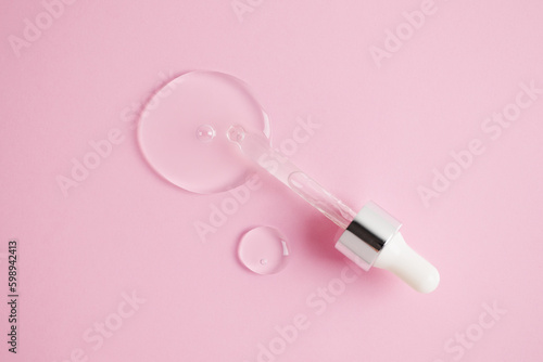 Glass pipette and drop of facial serum or oil on light pink background. Drop of liquid gel with hyaluronic acid closeup. Cosmetic product macro. Skin care with collagen. Beauty routine. Top view.