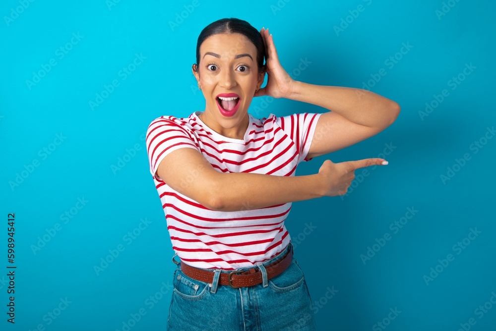 Naklejka premium beautiful woman wearing striped T-shirt over blue studio background feeling positive has amazed expression, indicates something. One hand on head and pointing with other hand.