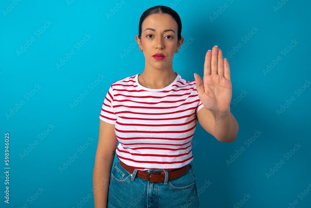 beautiful woman wearing striped T-shirt over blue studio background doing stop gesture with palm of the hand. Warning expression with negative and serious gesture on the face. beautiful woman wearing striped T-shirt over blue studio background doing stop gesture with palm of the hand. Warning expression with negative and serious gesture on the face.