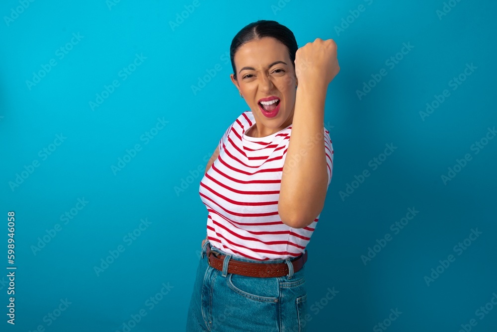 Overjoyed beautiful woman wearing striped T-shirt over blue studio background glad to receive good news, clenching fist and making winning gesture. Overjoyed beautiful woman wearing striped T-shirt over blue studio background glad to receive good news, clenching fist and making winning gesture.