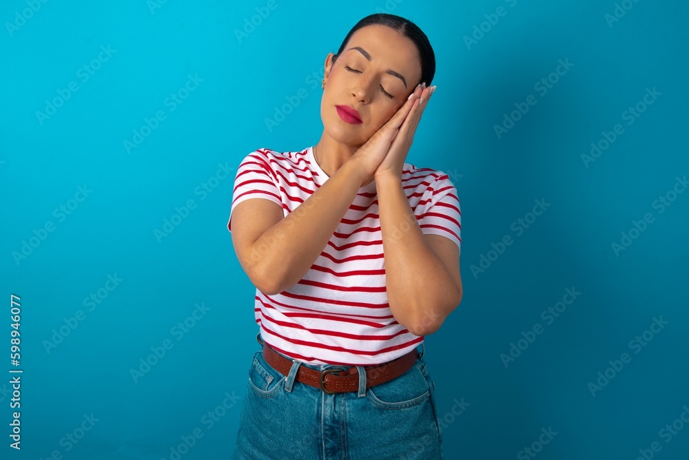 Relax and sleep time. Tired beautiful woman wearing striped T-shirt over blue studio background with closed eyes leaning on palms making sleeping gesture. Relax and sleep time. Tired beautiful woman wearing striped T-shirt over blue studio background with closed eyes leaning on palms making sleeping gesture.