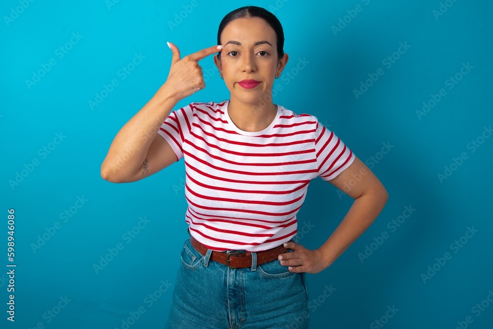 Naklejka premium Unhappy beautiful woman wearing striped T-shirt over blue studio background curves lips and makes suicide gesture, shoots in temple with hand, tries kill himself.