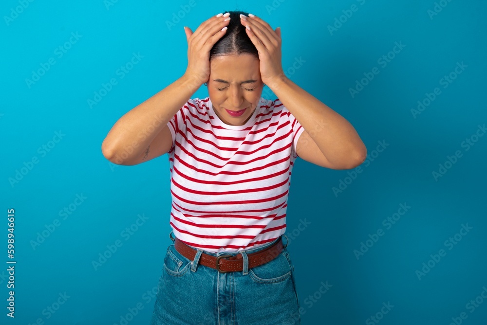 beautiful woman wearing striped T-shirt over blue studio background holding head with hands, suffering from severe headache, pressing fingers to temples beautiful woman wearing striped T-shirt over blue studio background holding head with hands, suffering from severe headache, pressing fingers to temples